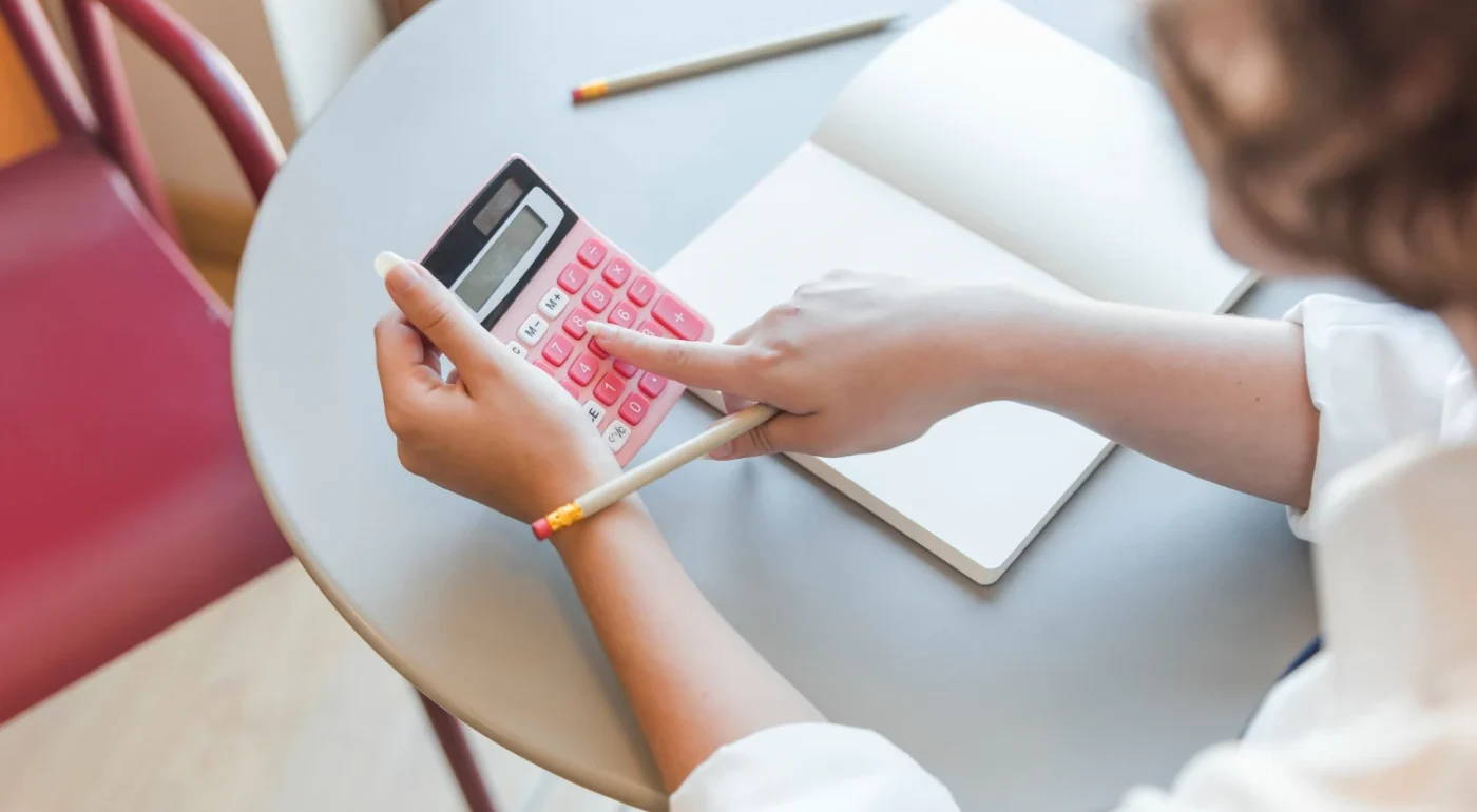 Pessoa usando uma calculadora rosa para administrar finanças pessoais com papel e lápis em uma mesa acomodada com cadeiras, ilustrando o Método do orçamento zero.